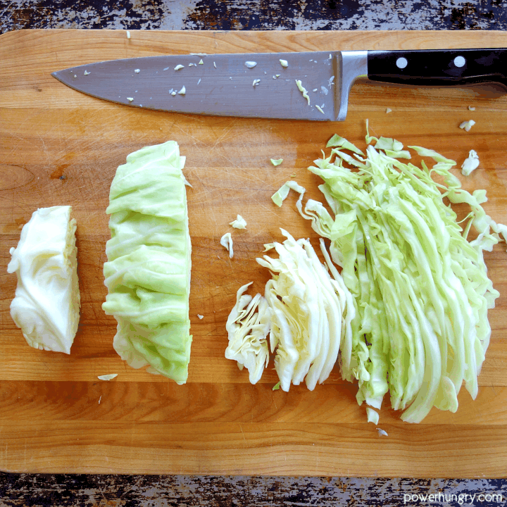 green cabbage on a wooden cutting board being cut into thin strips to make cabbage noodles