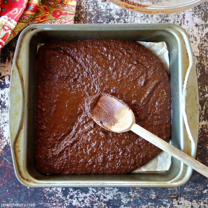 brownie batter spread into a parchment paper-lined square baking pan, a wooden spoon doing the spreading