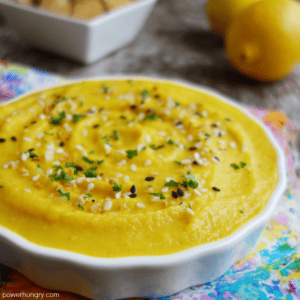 close up of lemon garlic split pea dip in a white bowl, with lemons and pita chips in the background