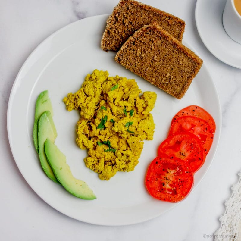 overhead shot of a white plate topped with vegan pumpkin seed scramble, tomatoes, avocado and whole grain bread.